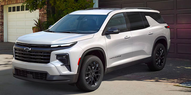 2025 Chevrolet Traverse SUV parked in a residential driveway, featuring a sleek white exterior, bold black grille, and stylish black alloy wheels with a modern, upscale design.