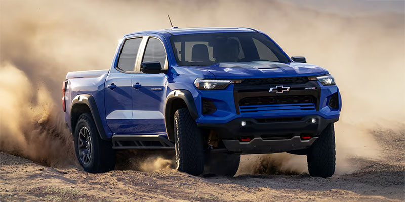 Front three-quarter view of a blue 2025 Chevy Colorado driving off-road through sandy terrain with dust clouds in the background.
