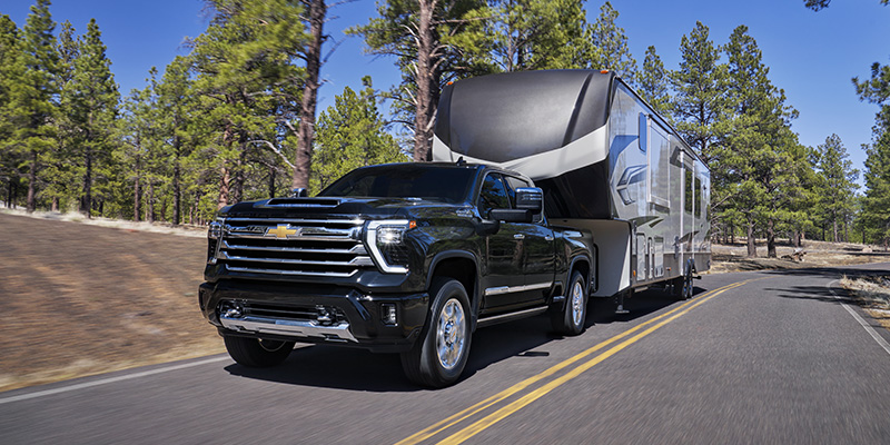 Black Chevrolet Silverado HD towing a fifth-wheel camper on a forest road.